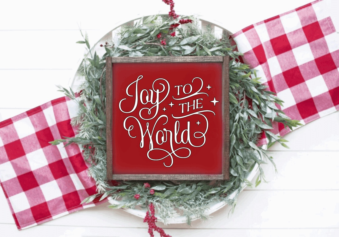 Joy to the World wood framed festive sign with red background and white lettering, surrounded by greenery and red plaid accents.