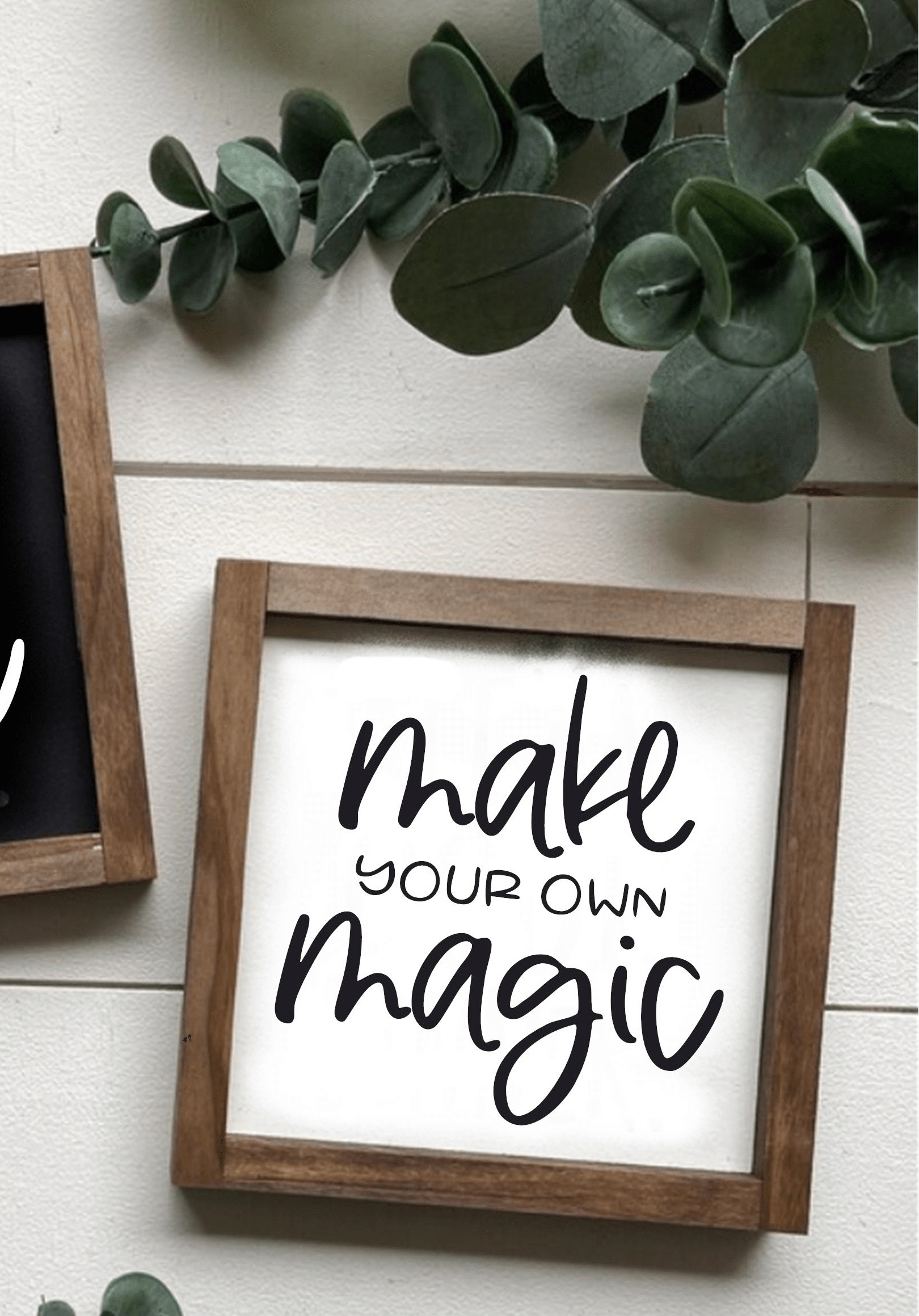 Hand-painted "Make Your Own Magic" wood sign with brown frame and white background, featuring black lettering, displayed against a white wall with greenery accents.