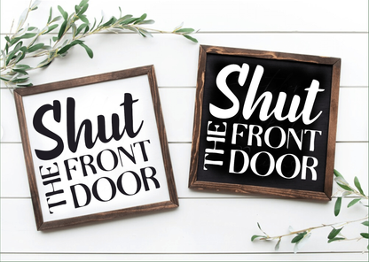 Handcrafted "Shut the Front Door" framed wood signs, black and white backgrounds with contrasting lettering, showcased on a white surface with greenery accents.
