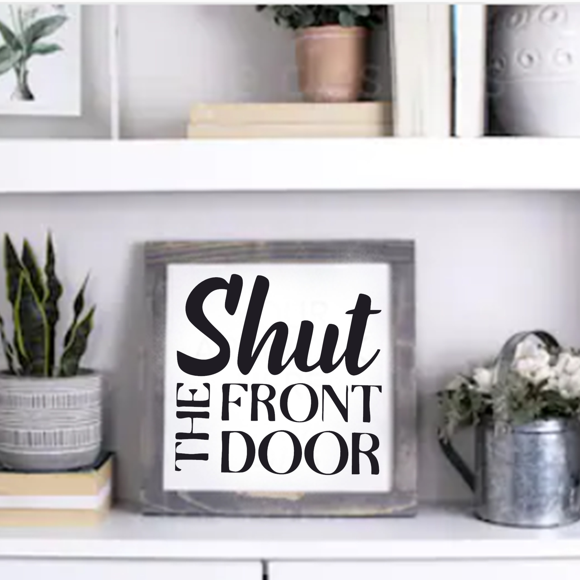 Shut the Front Door handcrafted framed wood sign with black lettering on white background, displayed on a shelf with plants and decor.