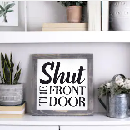 Shut the Front Door handcrafted framed wood sign with black lettering on white background, displayed on a shelf with plants and decor.