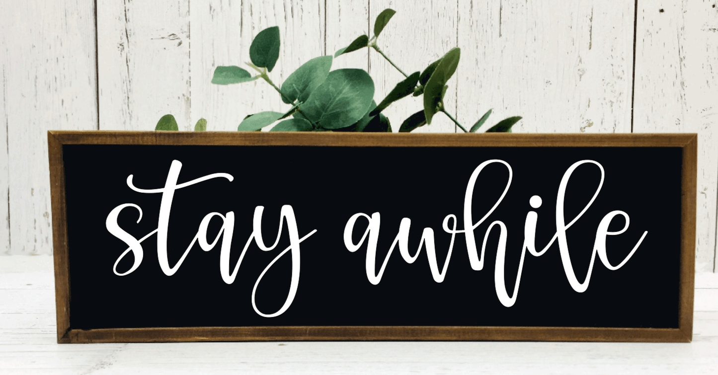 Handmade rustic wood sign reading "Stay Awhile" with black background and white lettering, framed in stained wood.