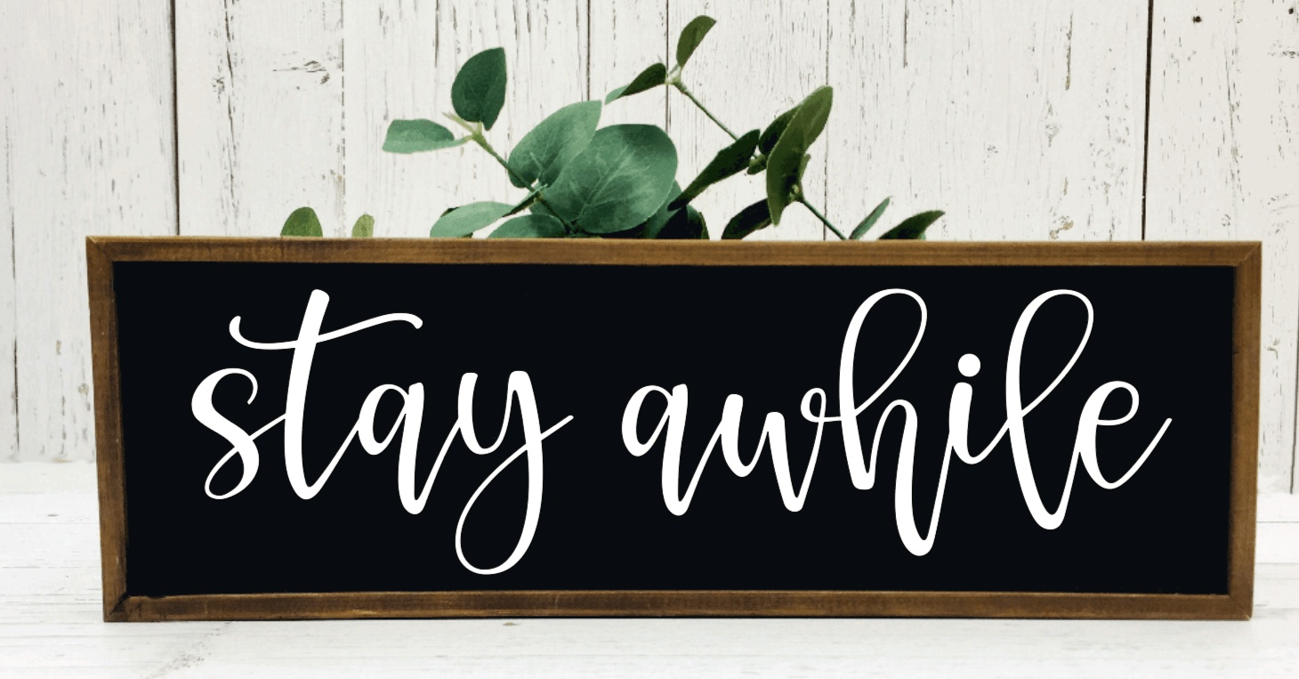 Handmade rustic wood sign reading "Stay Awhile" with black background and white lettering, framed in stained wood.
