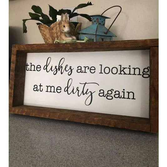 Rustic kitchen wood sign with the phrase "The Dishes are Looking at me Dirty Again" in black lettering on a white background, framed with pine wood.