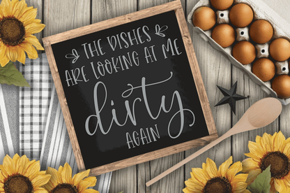 Funny kitchen sign saying "The Dishes are looking at me Dirty" with rustic wood frame on a table with sunflowers and eggs.