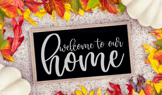 Framed wood sign with "Welcome To Our Home" in cursive on a black background, surrounded by autumn leaves.