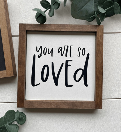 Hand-painted "You Are Loved" wooden sign with stained frame and white background featuring black lettering.