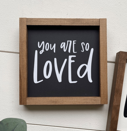 Inspirational "You Are Loved" sign with black background, white lettering, and wooden frame.