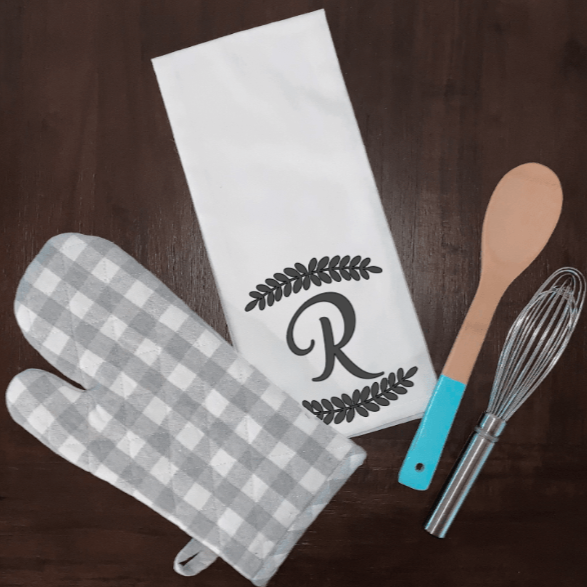 Personalized initial tea towel with leaf border, white microfiber, surrounded by kitchen utensils.