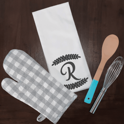 Personalized initial tea towel with leaf border, white microfiber, surrounded by kitchen utensils.