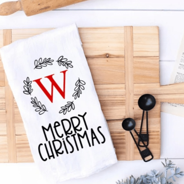 Personalized monogram Christmas tea towel with red initial and festive wreath design on a wooden board.