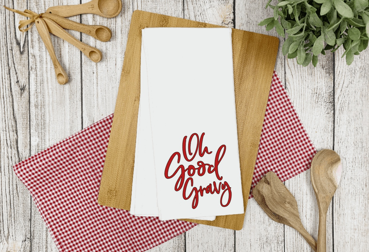 White tea towel with "Oh Good Gravy" saying, on wooden board with red checkered cloth.