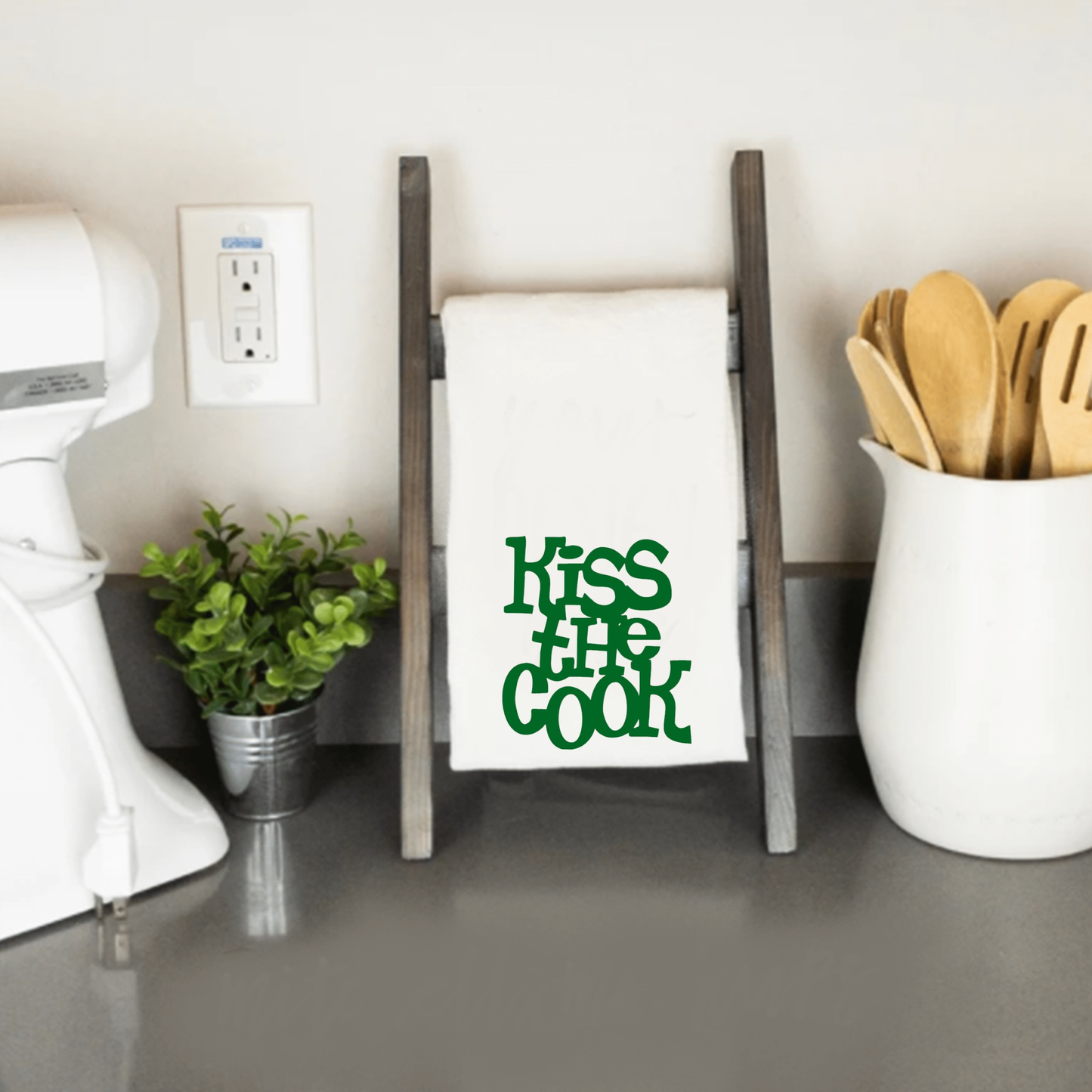 Humorous "Kiss the Cook" tea towel on a kitchen counter with utensils.