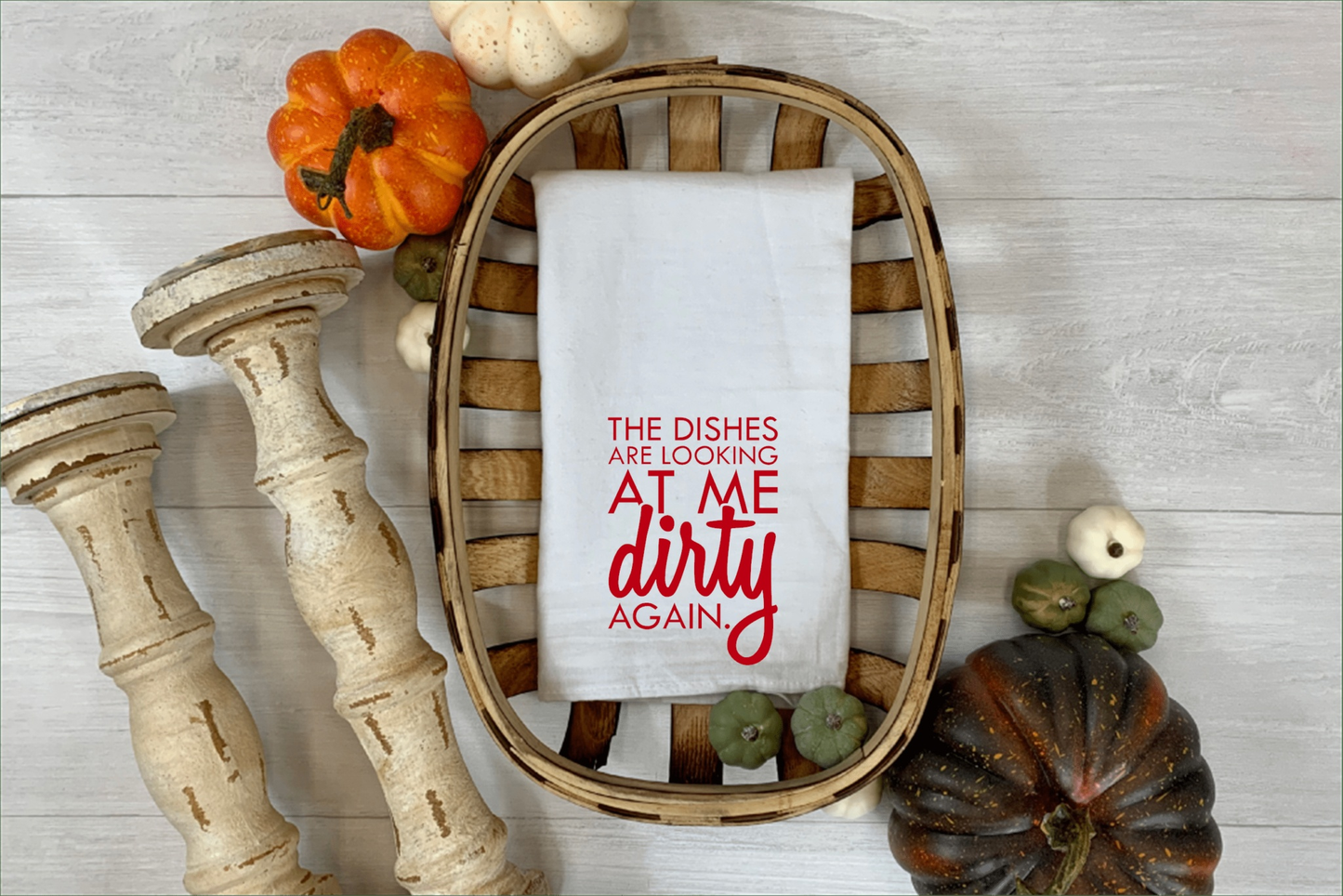 Funny "The Dishes are Looking at Me Dirty Again" white tea towel displayed in a decorative basket surrounded by pumpkins and candle holders.