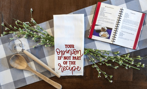 Decorative tea towel with humorous saying "Your Opinion is not part of the Recipe," white microfiber, waffle weave, surrounded by kitchen items.