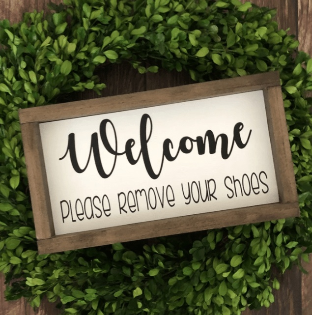 Handmade wood sign with "Welcome Please Remove Your Shoes" message, framed and set on a wreath display.