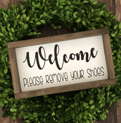 Handmade wood sign with "Welcome Please Remove Your Shoes" message, framed and set on a wreath display.
